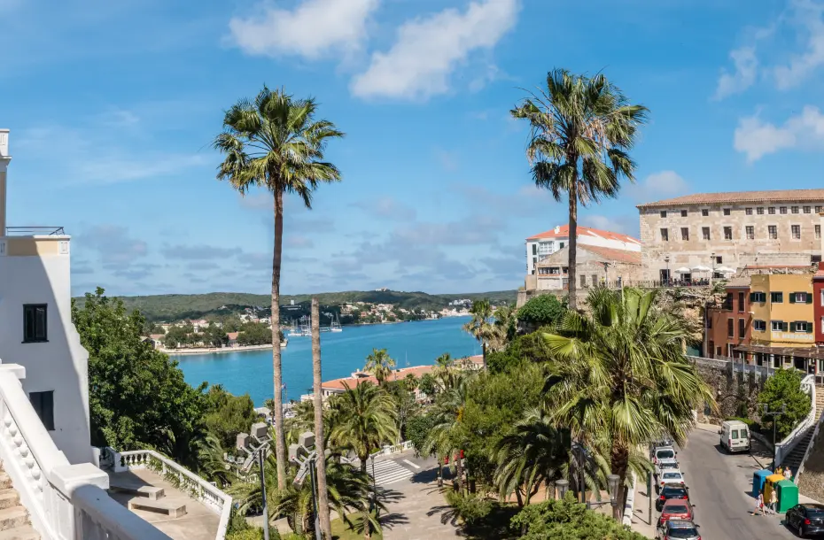 Vista del casco antiguo de Mahón y el puerto al fondo, detrás de altas palmeras.